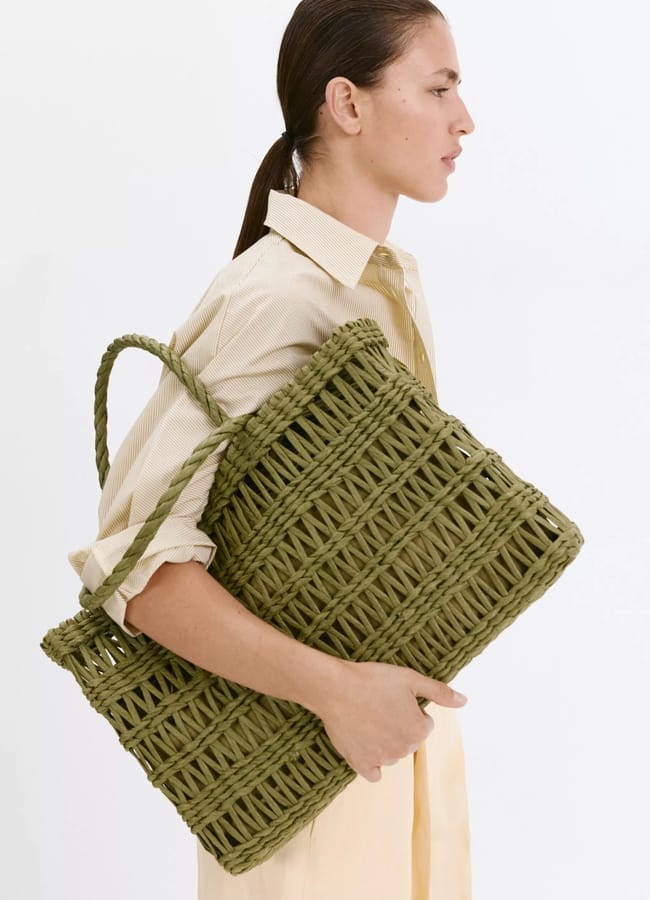 A woman with dark hair in a low ponytail wears a light yellow shirt dress—one of our top holiday fashion picks—and carries a large green woven tote bag under her arm against a plain white background.