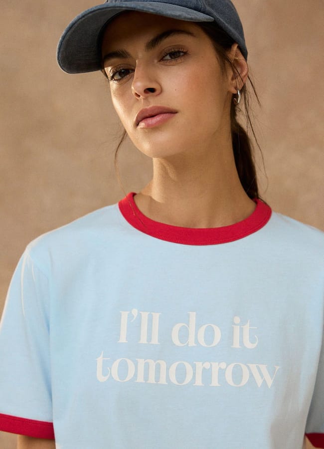 A woman wearing a blue cap and a light blue T-shirt with red trim that says “I’ll do it tomorrow” in white text stands against a beige background, showcasing one of this season’s best slogan shirts.
