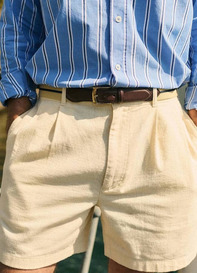A person wearing a blue and white striped button-up shirt tucked into cream-coloured shorts with hands in pockets and a brown belt around the waist, capturing refined men’s summer dressing inspired by The Talented Mr Ripley style.