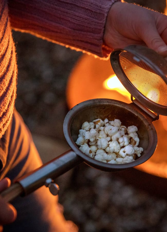 A person in a pink jumper holds an open popcorn maker with popped corn inside, sitting near an outdoor fire—perfectly capturing the cosy vibe of an outdoor film night at home.