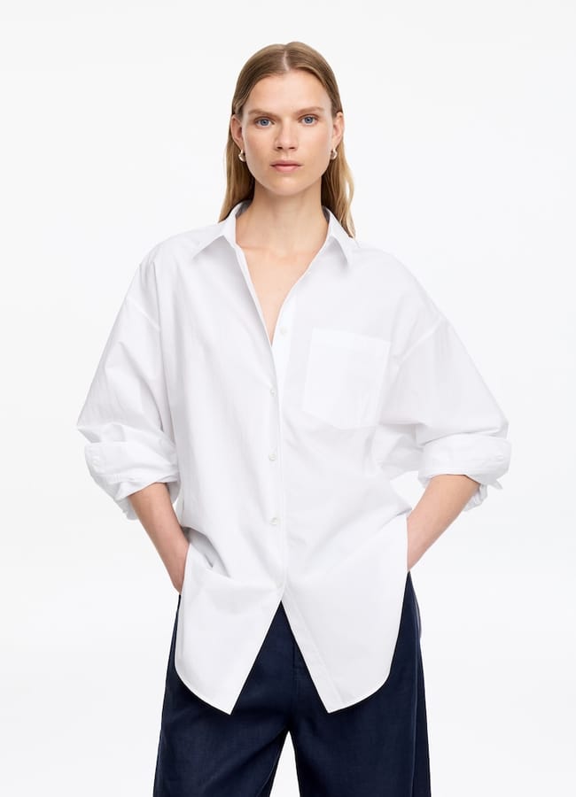 A person stands against a plain white background, wearing an oversized white button-down shirt with rolled-up sleeves and dark trousers—hands in pockets and a neutral expression—offering inspiration for effortless oversized shirt outfit ideas.
