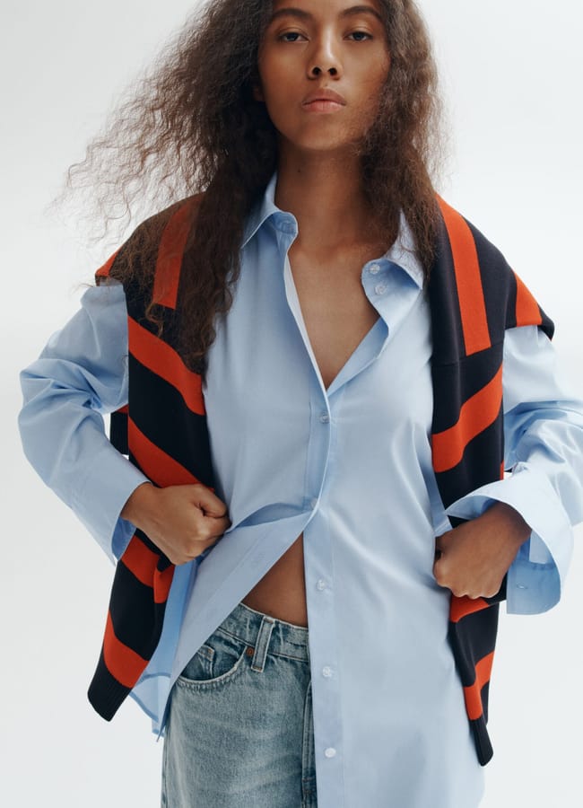 A person with long curly hair wears a light blue oversized shirt, partially unbuttoned, styled according to chic oversized shirt outfit ideas. A navy and red jumper is draped over their shoulders, paired with light blue jeans. They face the camera with a neutral expression.
