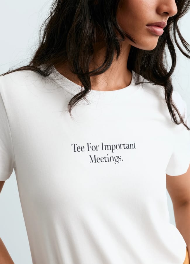 A woman with long dark hair wears a white T-shirt that has the text Tee For Important Meetings. printed in small black letters on the front. Only her shoulders, chest, and part of her face are visible, showcasing one of this season’s cool slogan tees.