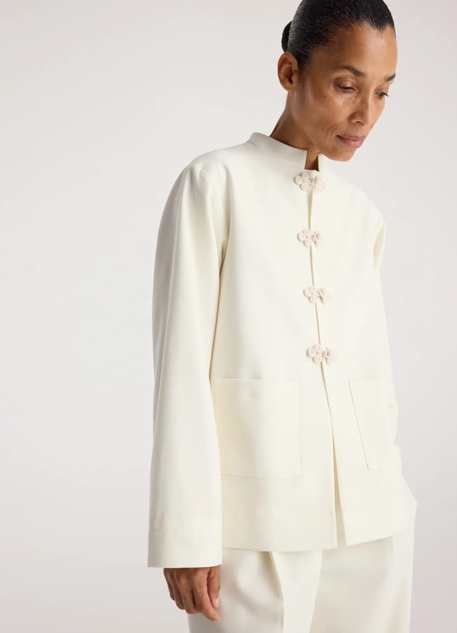 A person wearing an off-white, long-sleeved jacket with decorative knot buttons and large front pockets stands against a plain, light background—an effortless nod to autumn winter 2026 fashion trends.