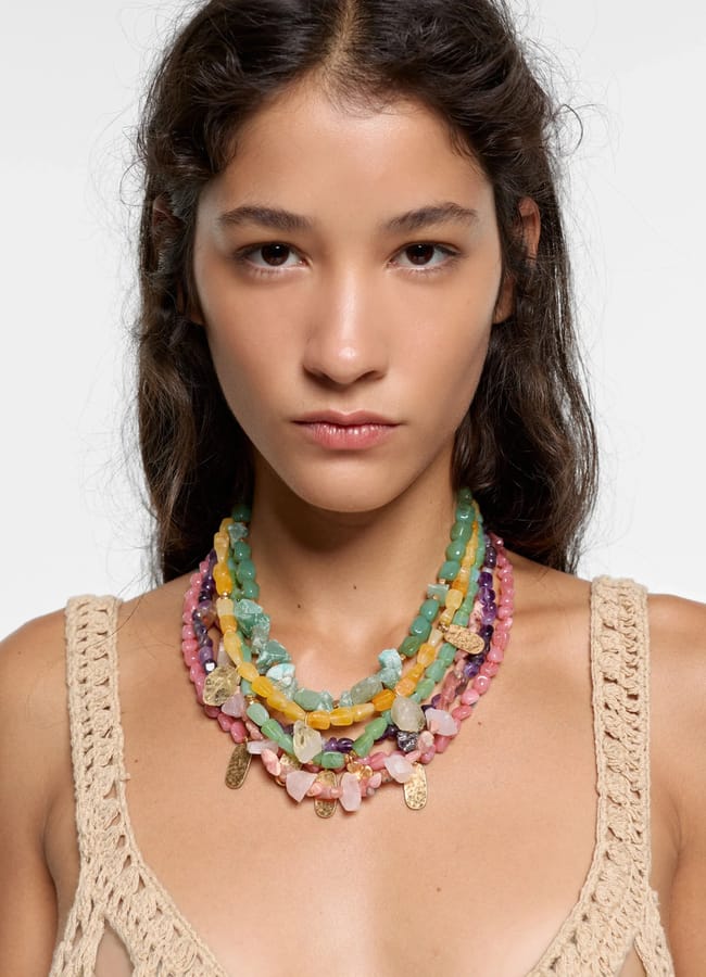 A woman with long brown hair wears a crocheted beige top and several colourful jewellery pieces—gemstone necklaces layered in shades of green, yellow, pink, and blue—against a plain light background.