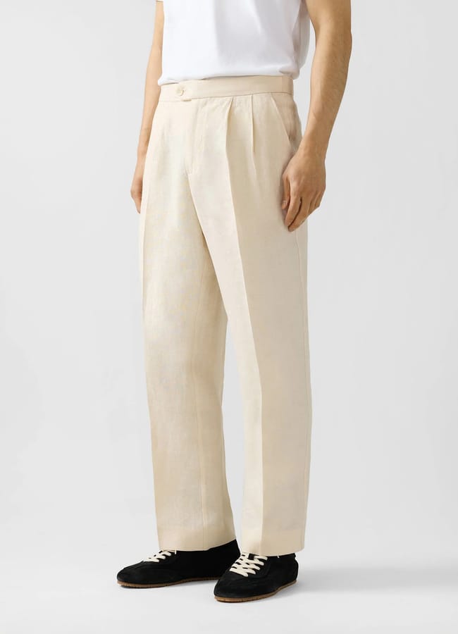 A person wearing a white T-shirt and beige pleated dress trousers with black trainers stands against a plain light background, channelling effortless men’s summer dressing inspired by The Talented Mr Ripley style.
