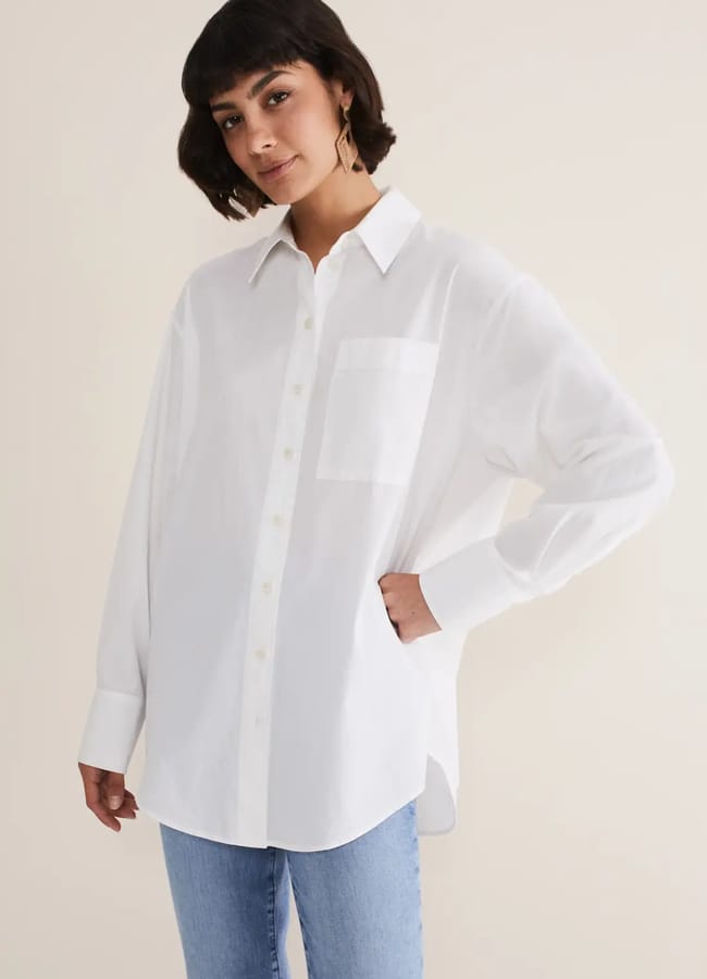 Woman wearing a loose white button-up shirt with a chest pocket, paired with light blue jeans, standing against a plain beige background—an effortless example for those seeking oversized shirt outfit ideas.