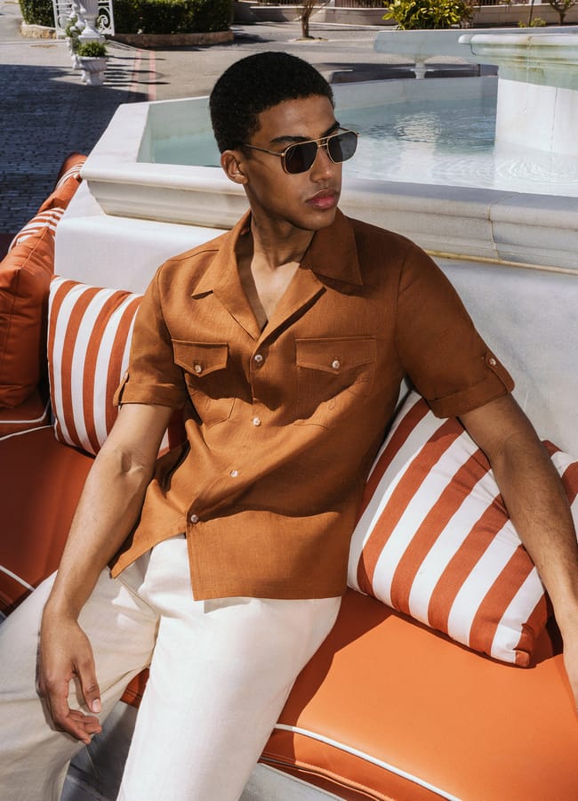 A man epitomising men’s summer dressing, wearing sunglasses, a brown short-sleeved shirt, and white trousers, sits on an orange and white striped sofa outdoors near a fountain—a look reminiscent of The Talented Mr Ripley style.