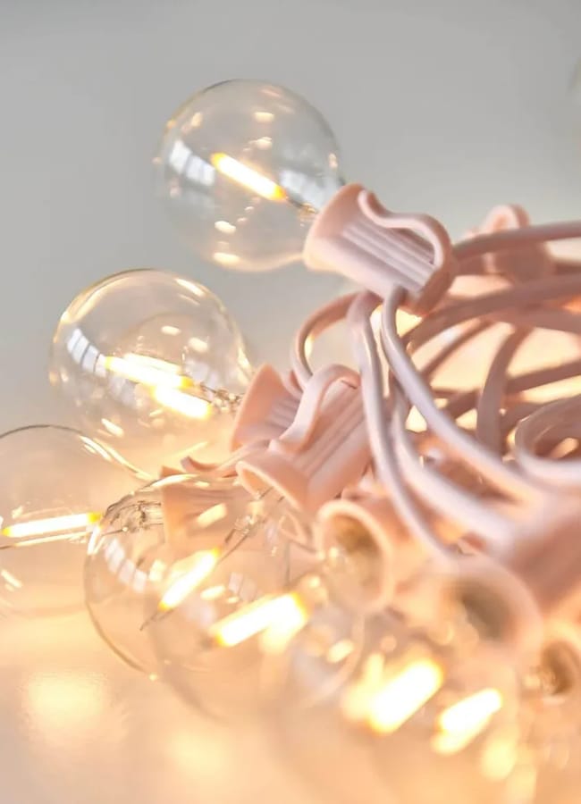 A close-up of glowing fairy lights with round glass bulbs and pink fittings, softly lit and coiled on a white surface—perfect for setting the mood to create an outdoor film night at home.