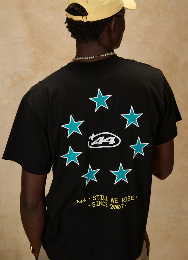 A person wearing a black T-shirt with teal stars and a +44 logo on the back, along with yellow text reading +44 STILL WE RISE SINCE 2007, exudes laid-back style reminiscent of celebrity couples on holiday. The ensemble is completed with a yellow cap and a beaded necklace.