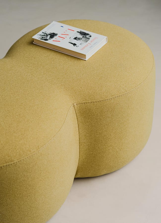 A beige, peanut-shaped ottoman sits on a light surface with a closed book featuring a mostly white cover and red text resting on top, subtly contrasting with the chartreuse colour trend that’s currently making waves in modern interiors.