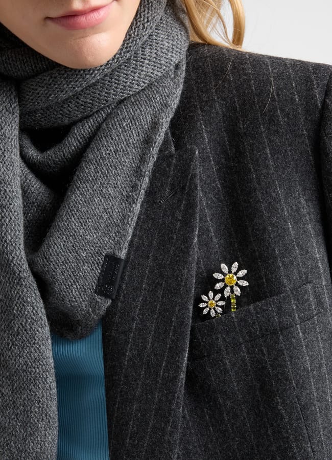 A person in a grey pinstripe blazer and grey scarf, perfectly embodying autumn winter 2026 fashion trends, has a daisy-shaped brooch with yellow and white stones on their lapel. Only the lower half of their face is visible.