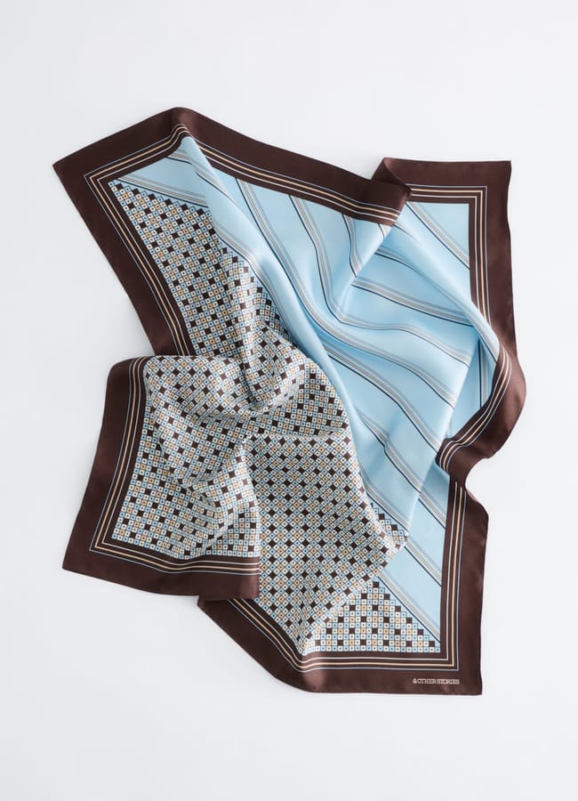 A silk scarf with a brown border, featuring a blue section with thin stripes and a section with a detailed geometric pattern in brown, white, and blue, is laid flat but appears gently gathered in the centre—a perfect accessory to elevate your holiday fashion and make those holiday photos truly stand out.