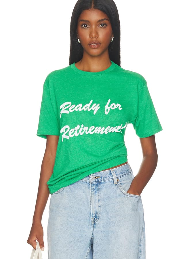 A woman wearing a green T-shirt with the cool slogan tee “Ready for Retirement” printed in white letters, paired with light blue jeans. She has long dark hair and is holding a light-coloured bag.
