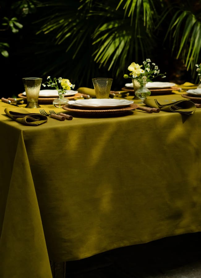 A yellow tablecloth covers a table set with plates, glasses, and small vases of yellow flowers, subtly echoing the chartreuse colour trend. The setting is outdoors, with green palm leaves in the background.