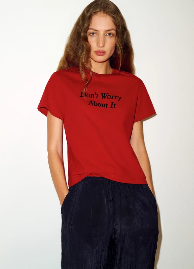Woman with long wavy hair wearing a red slogan T-shirt that says Don’t Worry About It in black text, paired with loose-fitting black trousers, standing against a plain light background.