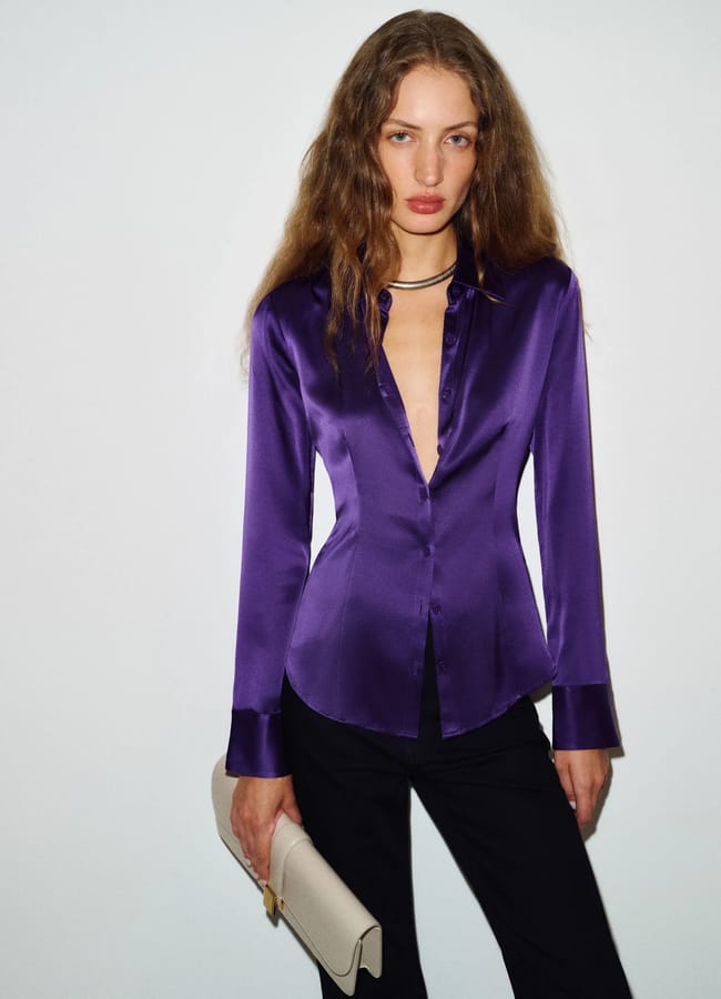 A woman with long wavy hair wears a fitted, shiny purple button-up shirt and black trousers, holding a beige clutch, standing against a plain white background—a chic look that perfectly captures autumn winter 2026 fashion trends.