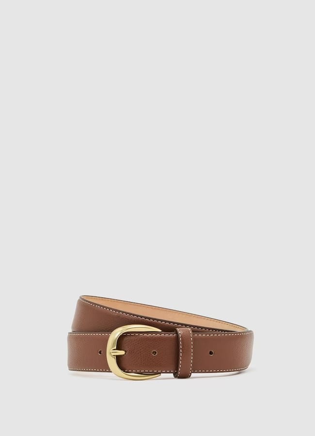 A brown leather belt with white stitching and a gold oval buckle is coiled against a plain white background, evoking the effortless elegance of Talented Mr Ripley style—an impeccable choice for elevating men’s summer dressing.