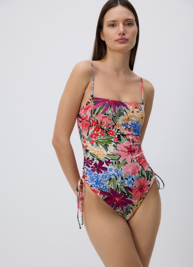 A woman wearing pretty swimwear—a colourful, floral one-piece swimming costume with thin straps and side ties—stands against a plain light background, looking at the camera with a relaxed expression.