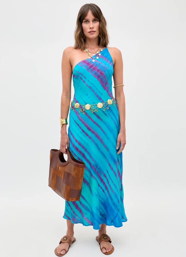 Woman wearing a blue and purple one-shoulder dress inspired by turquoise fashion, with gold accessories, brown sandals, and holding a brown bag, standing against a plain light grey background.