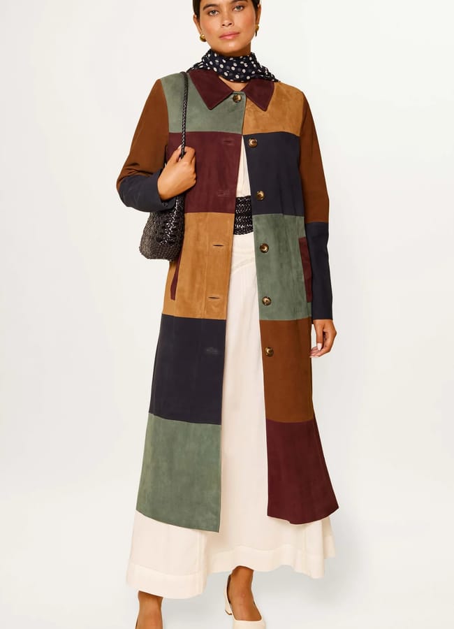 A woman wears a long, colour-block patchwork coat with earthy tones over a cream skirt, polka dot scarf, and cream shoes—an effortlessly chic look perfect for the summer sale season. She holds a textured bag and poses against a plain background.