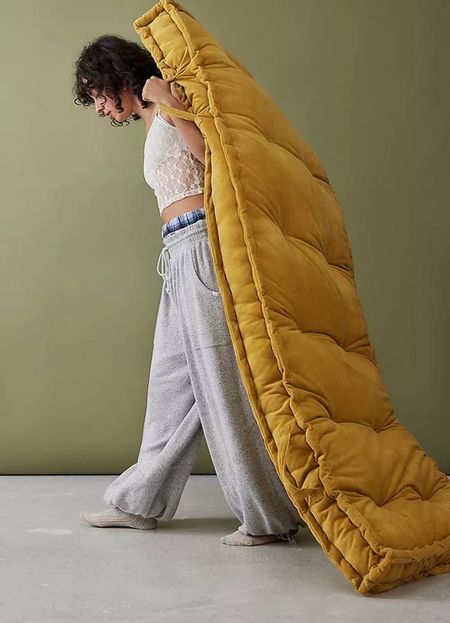 A person in a lace top and grey sweatpants carries a large, mustard-yellow quilted blanket over their shoulder, walking indoors against an olive-green wall—perhaps preparing to create an outdoor film night or cosy at home film night.
