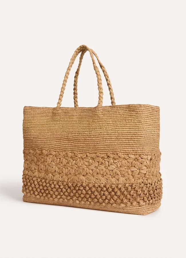 A large woven straw tote bag with two plaited handles, perfect for complementing your oversized shirt outfit ideas. The bag features textured, decorative patterns near the base and a smooth weave on the upper section.