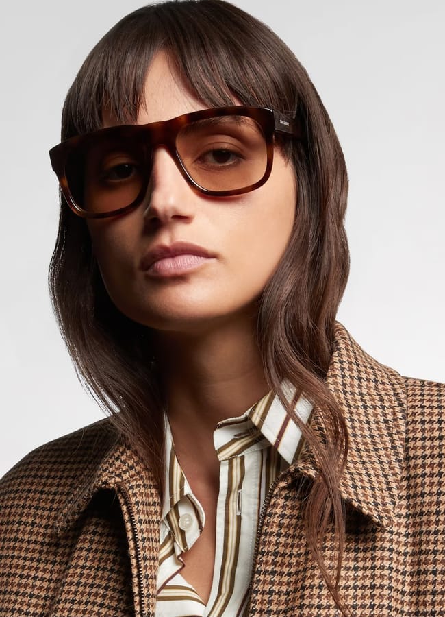 A woman with long brown hair wears large brown sunglasses, a brown houndstooth coat, and a striped shirt, posing against a plain light background—perfectly styled for the summer sale season.