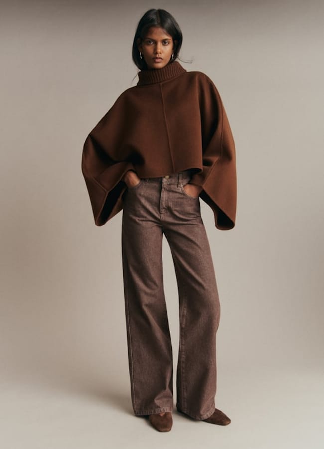 Woman stands against a beige background wearing a brown oversized polo-neck poncho and wide-leg brown trousers with matching shoes, posing with hands in pockets—a sophisticated ensemble that perfectly captures the essence of autumn winter 2026 fashion trends.