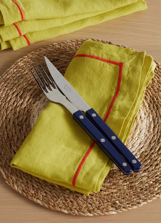 A fork and knife with blue handles rest on a yellow serviette with red trim, placed on a woven straw table mat atop a wooden table—a striking arrangement that echoes the chartreuse colour trend. Another folded yellow serviette sits in the corner.