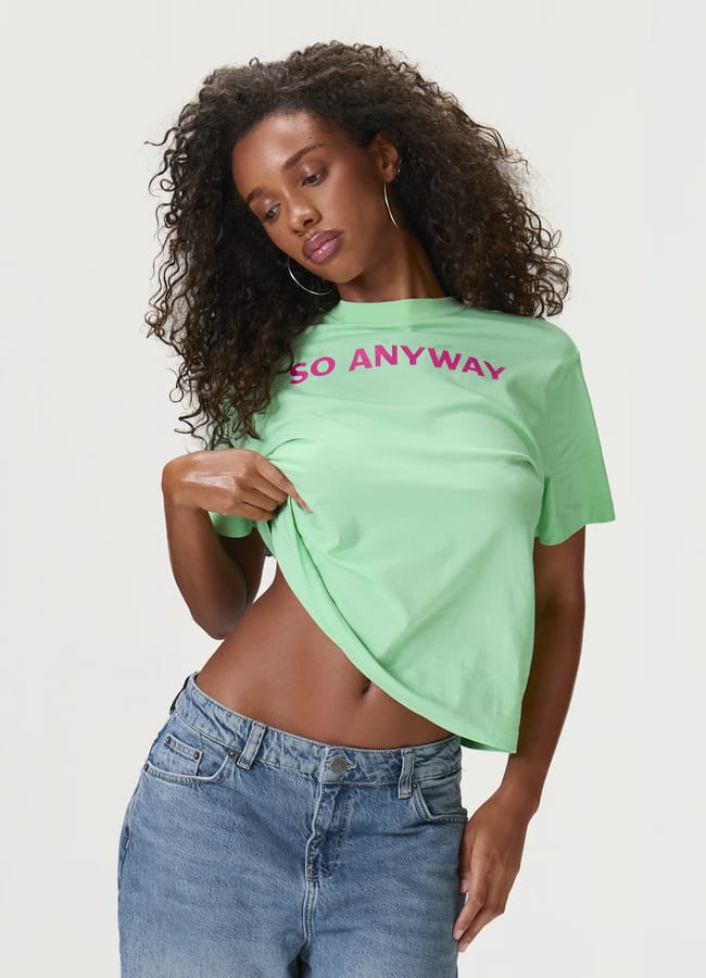 A woman with curly hair wears hoop earrings, a green cool slogan T-shirt with SO ANYWAY in pink letters, and blue jeans. She lifts one side of her shirt, exposing her waist, and looks downwards. The background is plain white.