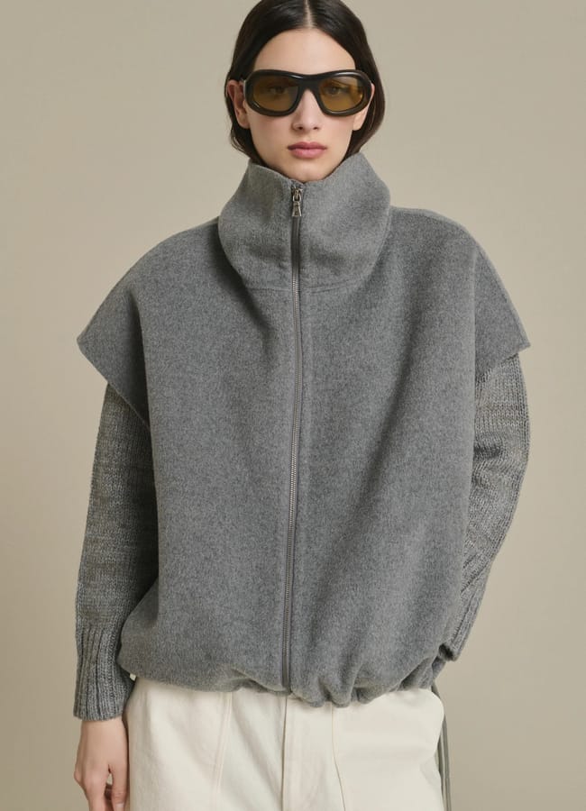 Woman wearing oversized sunglasses, a grey zip-up high-neck jumper layered over a knitted top, and off-white trousers, standing against a plain beige background—an effortlessly chic look inspired by autumn winter 2026 fashion trends.
