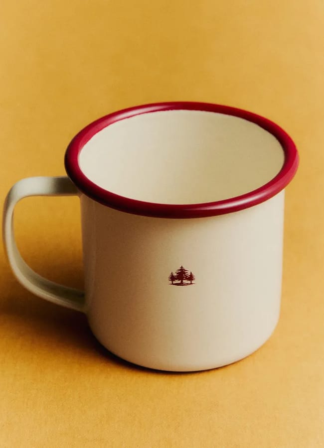 A cream-coloured metal mug with a red rim and a small tree design on the side sits on a plain light brown surface, ready for your cosy outdoor-film-night-at-home.
