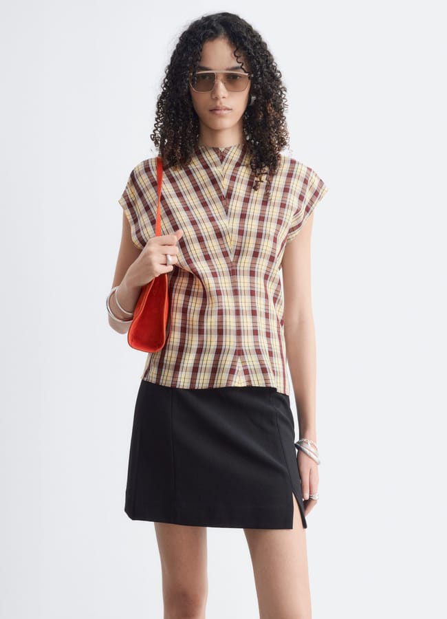 A woman with curly hair wears sunglasses, a checked top, a black mini skirt with a slit, silver bangles, and carries a red shoulder bag, standing against a plain white background—effortlessly showcasing autumn winter 2026 fashion trends.