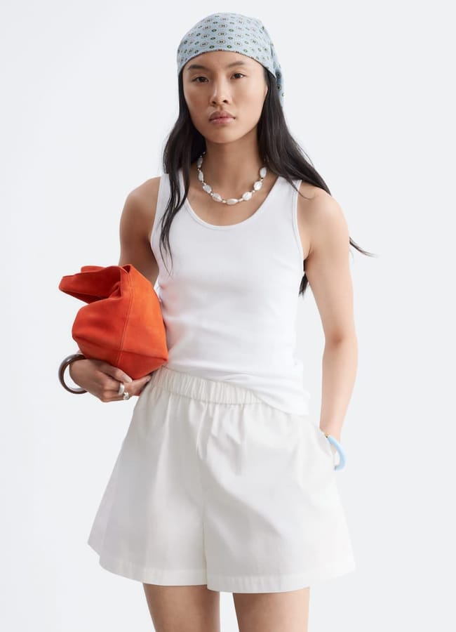 A woman wearing a white vest, white shorts, and a patterned headscarf holds an orange bag—a perfect look for those seeking last minute holiday style inspiration. She accessorises with a shell necklace and silver bracelet, posing against a plain light background for effortlessly chic holiday fashion pics.