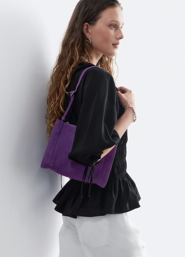 A woman with long wavy hair wearing a black top and white trousers holds a purple shoulder bag while looking to the side against a plain, light background, effortlessly showcasing one of the standout autumn winter 2026 fashion trends.