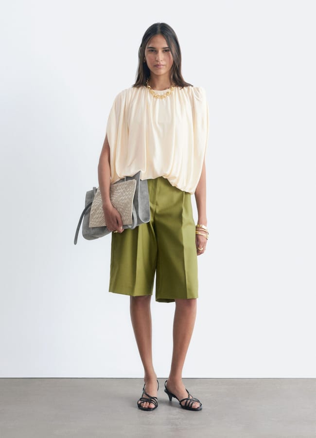A woman stands against a white background, wearing a cream blouse, olive green knee-length shorts, black sandals, and holding a large grey handbag—an effortlessly chic look that’s perfect for last minute holiday fashion. She accessorises with gold bracelets and a necklace, making this outfit ideal for spontaneous holiday fashion photos.