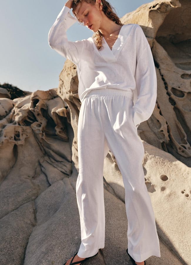 A person in a loose white outfit and black sandals stands on rocky terrain, with one hand on their head, looking down, as if savouring the spontaneous freedom of a last minute holiday, against a clear blue sky.