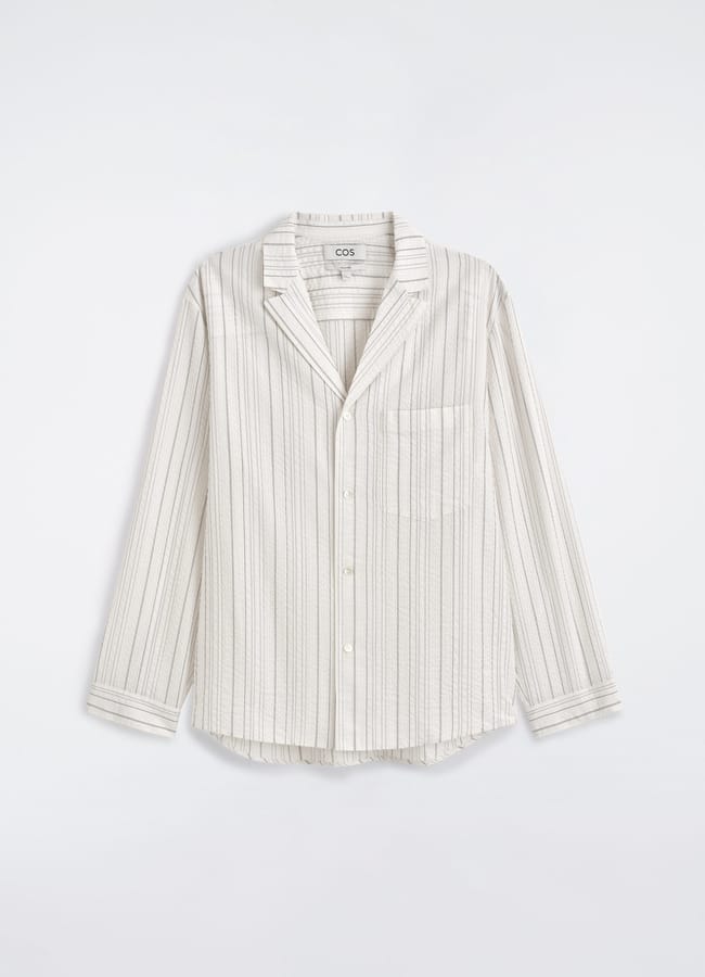A long-sleeve, button-up shirt with vertical beige and white stripes, a chest pocket, and a collar, displayed on a plain light background—perfect for recreating the effortless style often seen on celebrity couples on holiday.