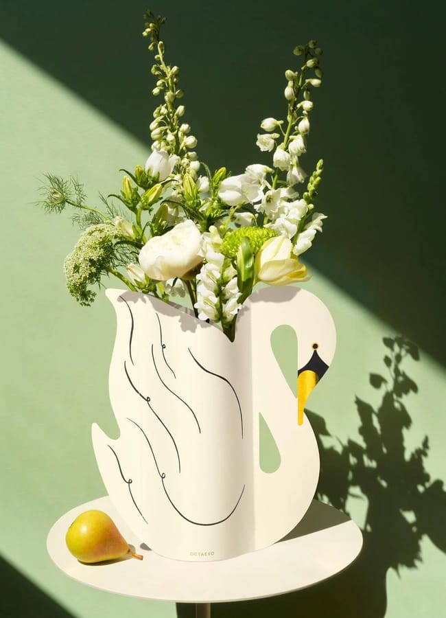 A swan-shaped white statement vase holding white and yellow flowers sits on a round table, with a yellow pear nearby, all illuminated by sunlight against a green background.
