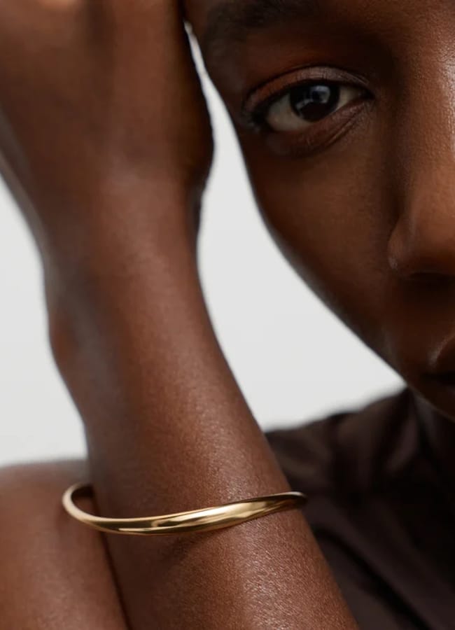 Close-up of a person with dark skin wearing a simple gold bracelet on their arm, resting their head on their hand. The background is plain and light-coloured, offering inspiration for oversized shirt outfit ideas.