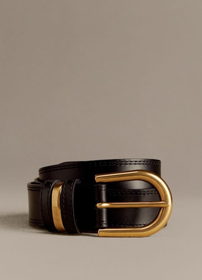 A rolled black leather belt with a shiny gold buckle and gold belt loop, displayed against a plain beige background—it’s the kind of timeless accessory you might spot styled effortlessly by celebrity couples on holiday.