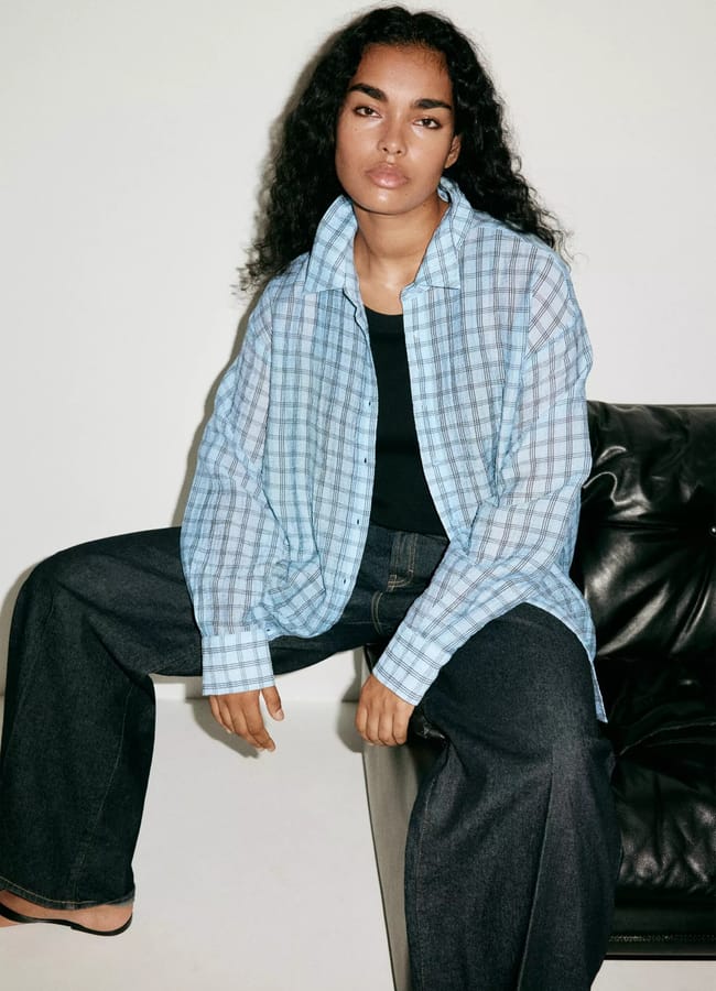 A person with long curly hair sits on a black sofa, wearing a loose light blue checked shirt over a black top and wide-leg dark jeans, looking confidently at the camera—perfect inspiration for oversized shirt outfit ideas.