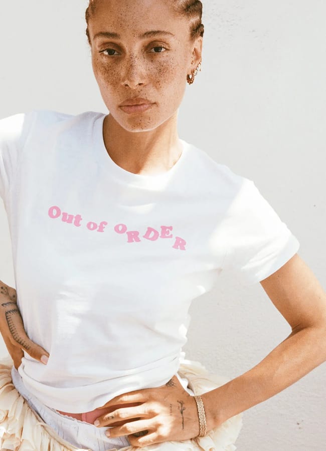 A person stands against a white wall, wearing one of those cool slogan tees—a white T-shirt with pink text that reads Out of ORDER. They have plaited hair, freckles, and a neutral expression.