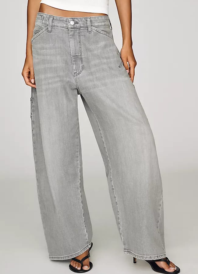 Person wearing wide-leg, light grey jeans and black heeled sandals, showcasing a stylish take on oversized shirt outfit ideas. Only the lower half of the body is visible, with one hand resting on the thigh. Background is plain and light-coloured.