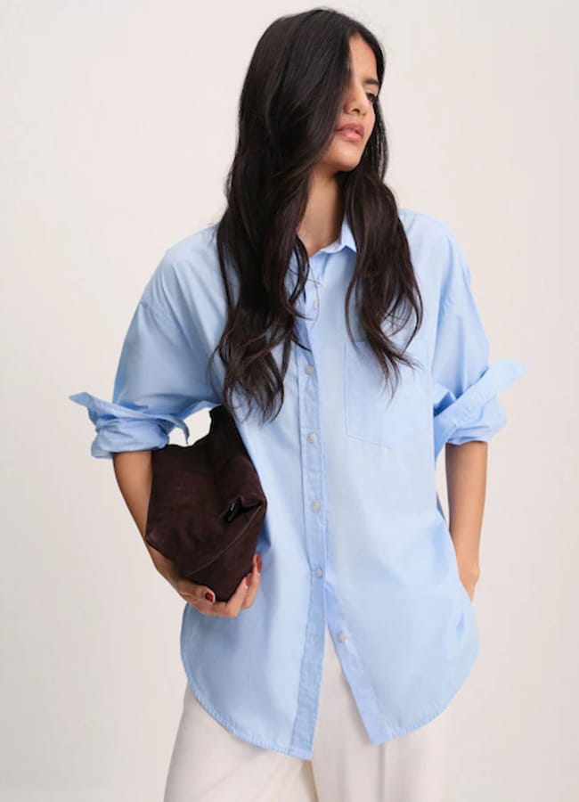 A woman with long dark hair wears a loose light blue button-up shirt and white trousers, holding a dark brown clutch under her arm, standing against a plain light background—offering chic inspiration for oversized shirt outfit ideas.