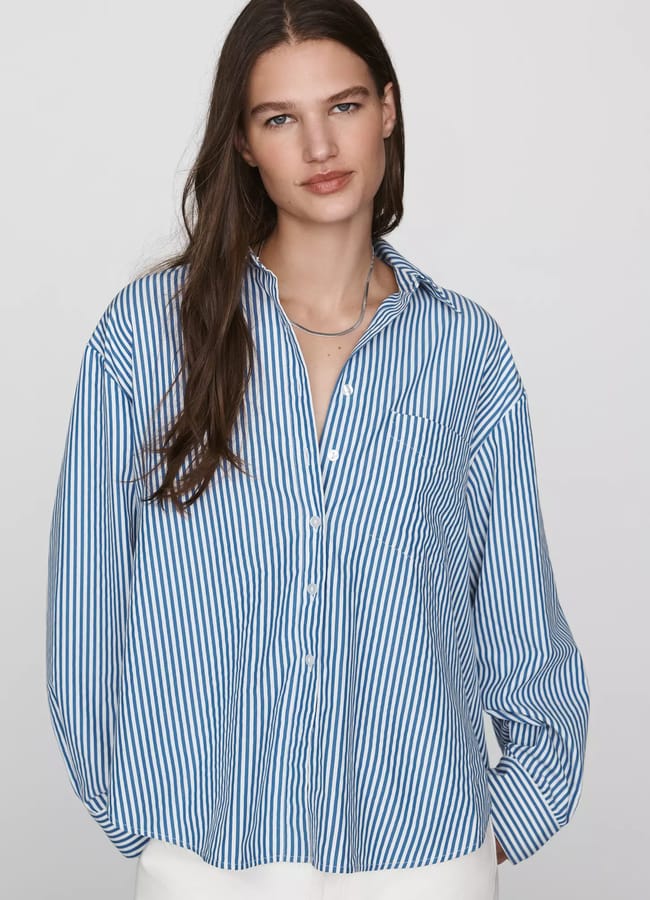 Woman with long brown hair wearing a loose blue-and-white striped button-up shirt and white trousers, standing against a plain light grey background. Showcasing one of the season’s best oversized shirt outfit ideas, she has a neutral expression and a slight head tilt.