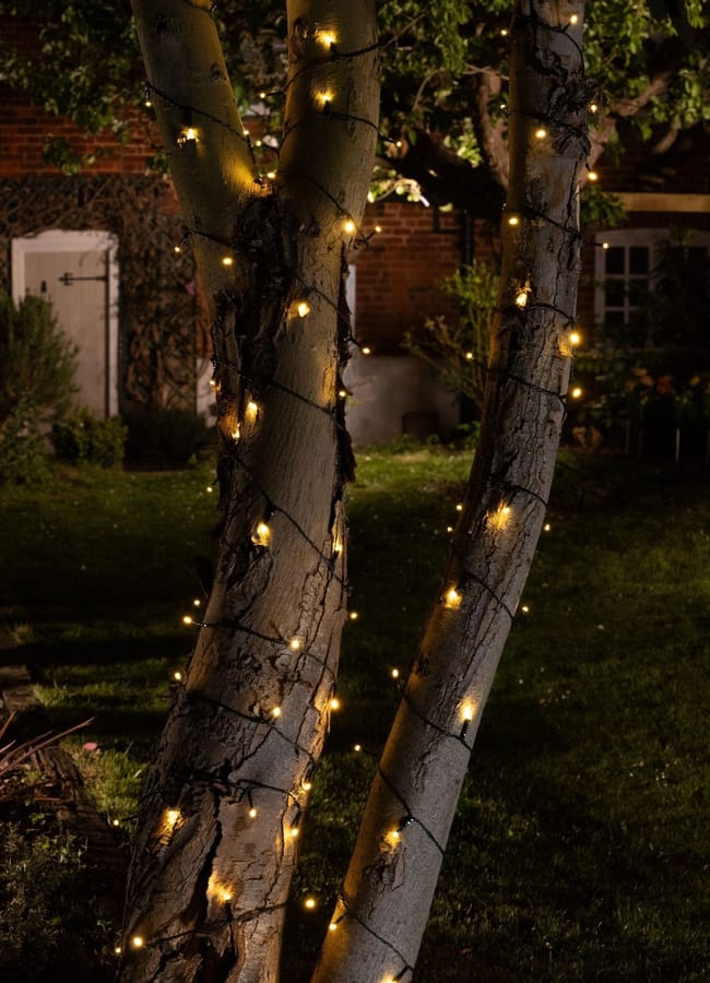 Two tree trunks wrapped with small warm white fairy lights illuminate a garden at night, setting the perfect ambience to create an outdoor film experience. A brick house with a white door and lush garden plants form a cosy backdrop, ideal for an unforgettable at-home film night.
