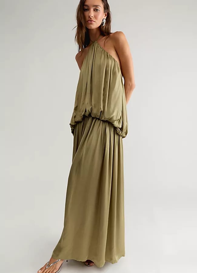 A woman in a loose, olive green halter-neck maxi dress and sandals stands in a minimal studio setting, looking at the camera with a relaxed pose—perfectly capturing effortless holiday style inspired by celebrity fashion.