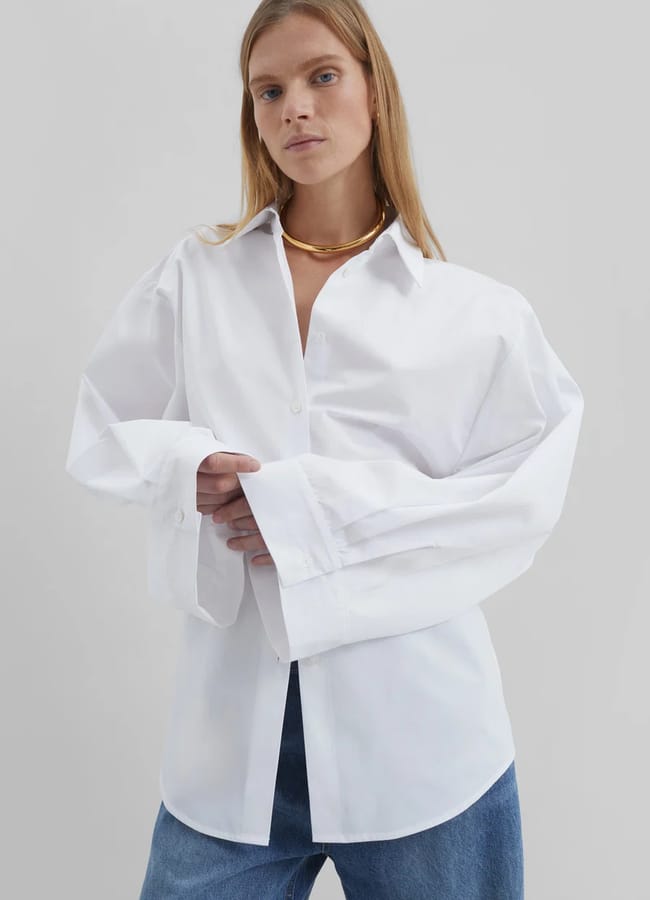 A person wearing an oversized white button-up shirt with long sleeves and blue jeans stands against a plain grey background, showcasing one of the classic oversized shirt outfit ideas.